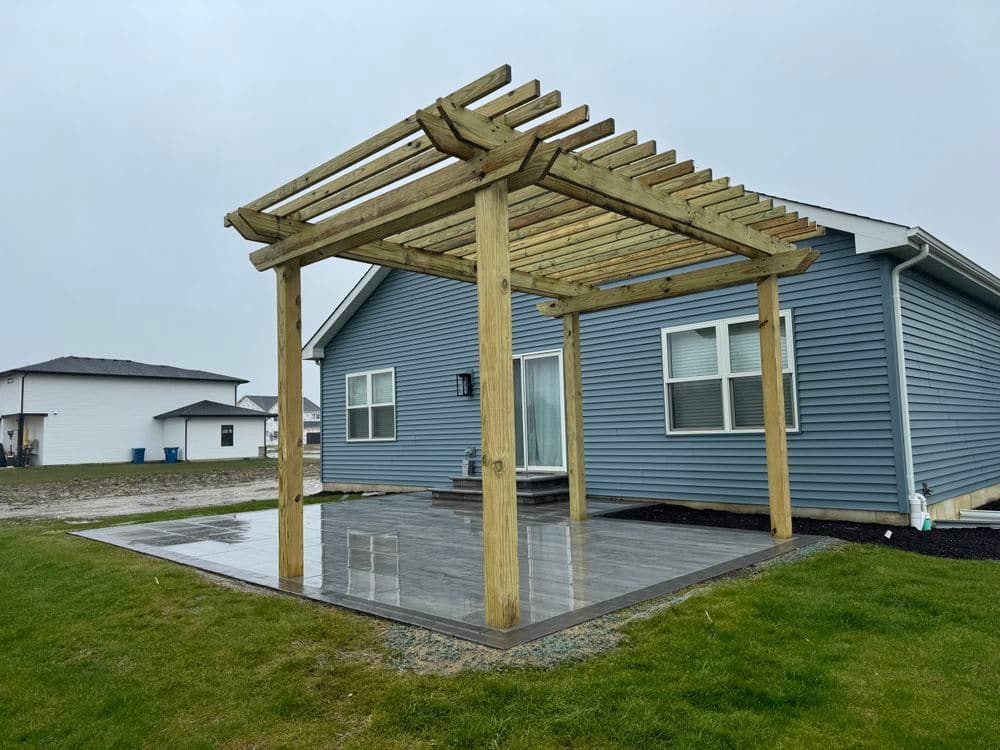 Wooden pergola over a stone patio in front of a blue house on a rainy day.