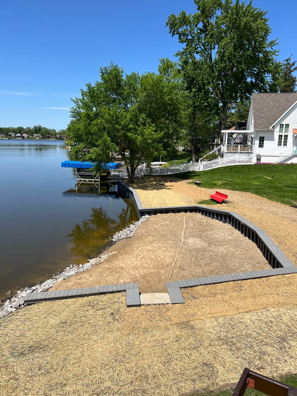 Lakeside view with sandy area, dock, and house under blue sky and greenery.