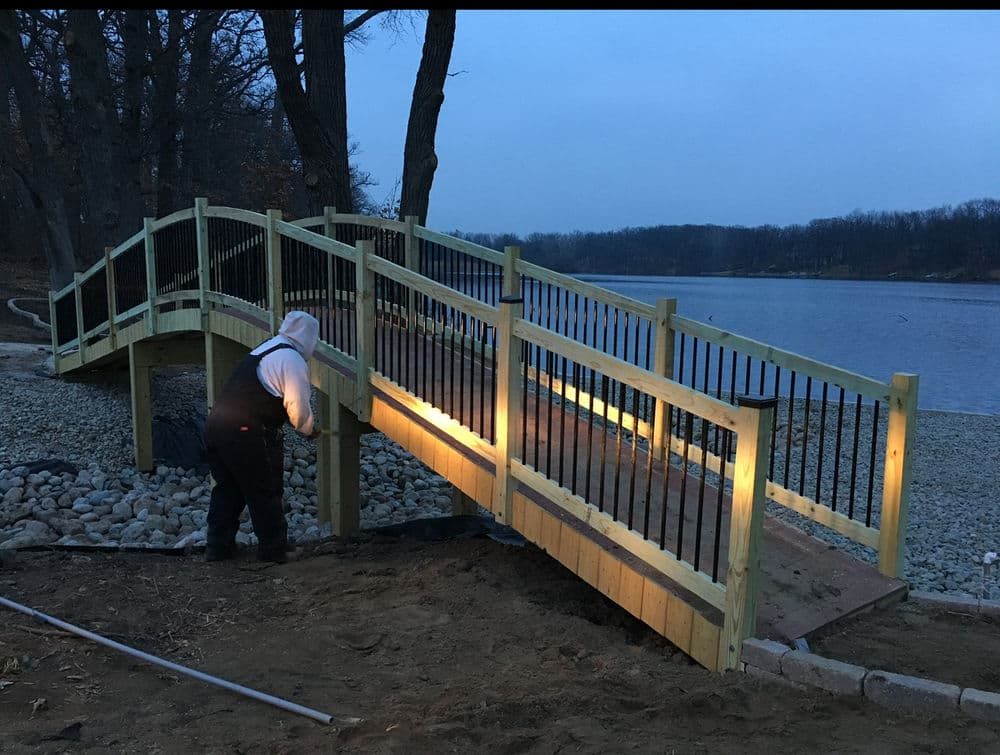 Wooden bridge under construction by a river at dusk, featuring guardrails and lighting.